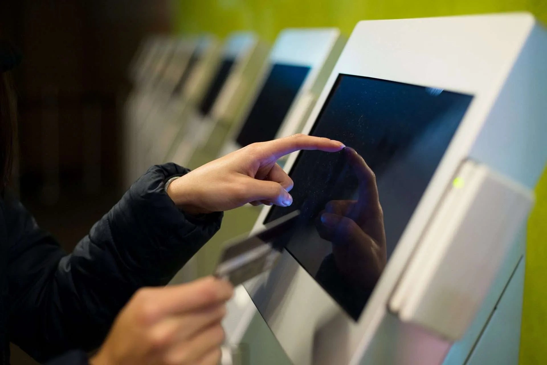 Person's hand touching a dark touchscreen monitor on a white kiosk, with a credit card in the other hand.