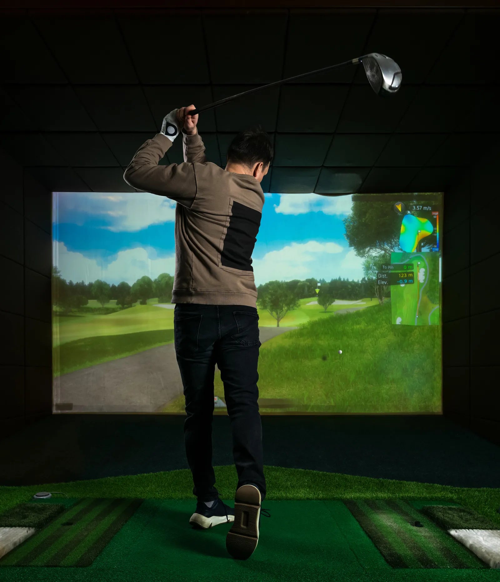 Man in a brown sweater and jeans swinging a golf club in a realistic 4K golf simulator.
