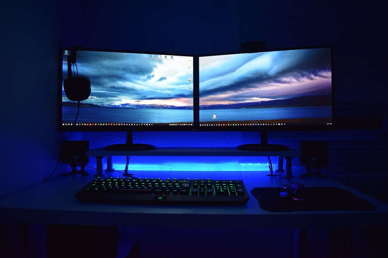 Dual monitors displaying a scenic landscape, illuminated by blue LED backlighting, on a desk with a gaming keyboard and mouse.