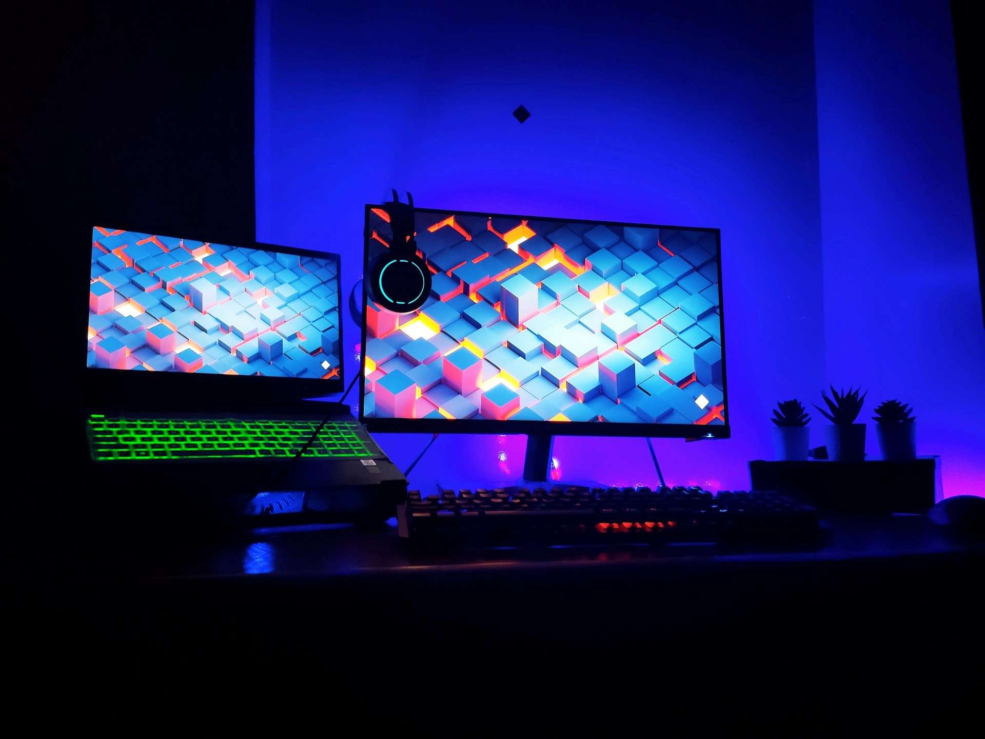 Gaming setup with a laptop and monitor displaying a vibrant geometric pattern, illuminated by blue and green RGB lighting.