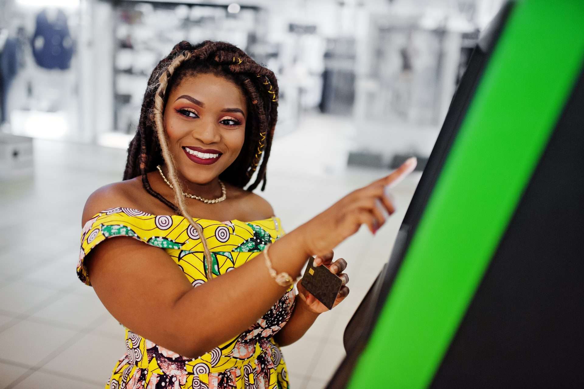 Smiling woman in a vibrant yellow dress using a green-screen self-ordering kiosk with a credit card.