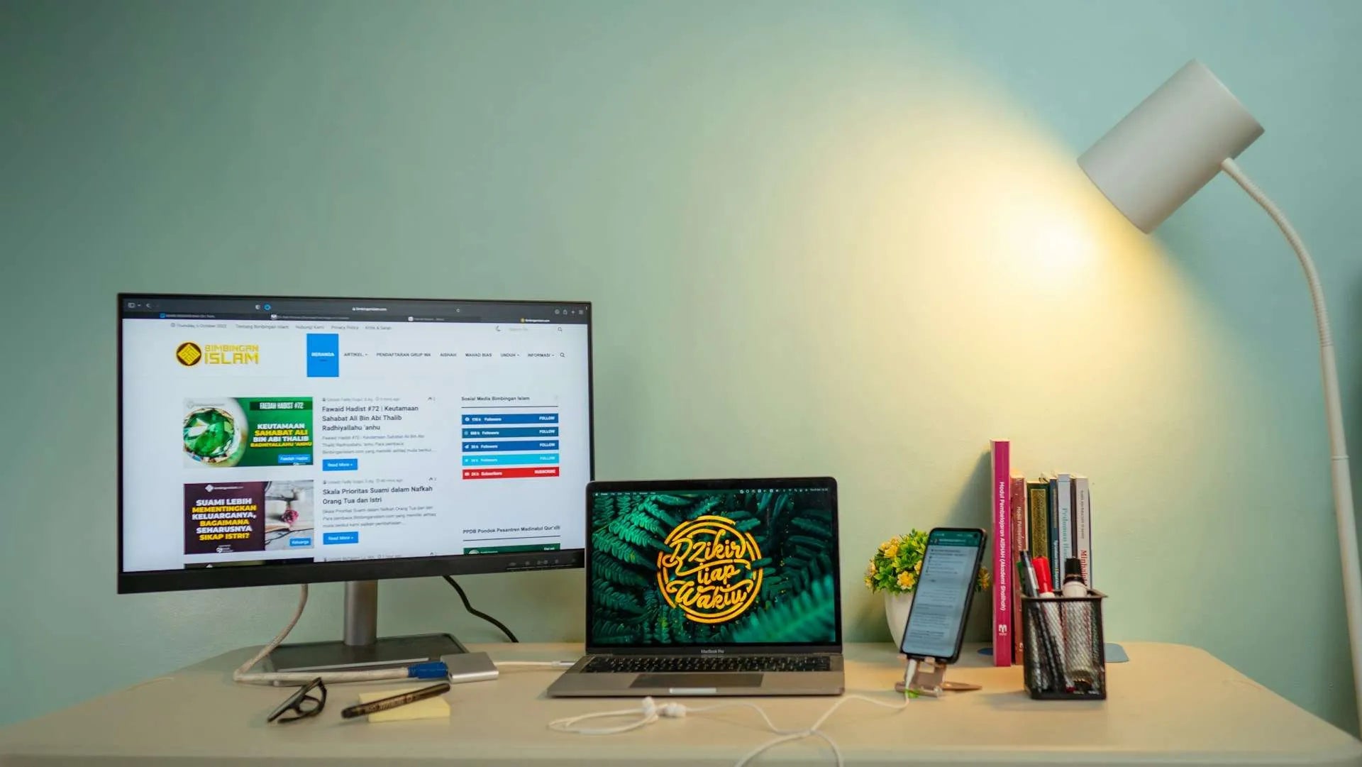A modern desk setup with a monitor, laptop, smartphone, and books, illuminated by a white desk lamp.