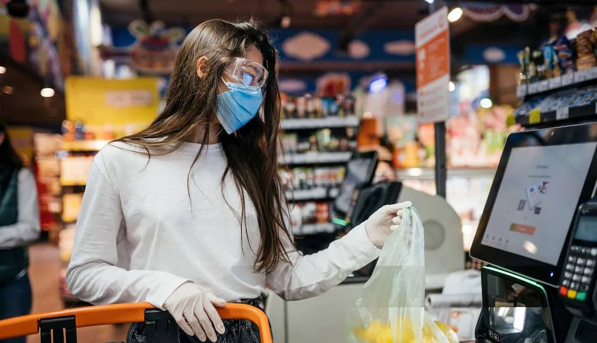 Woman in mask, goggles, and gloves using a self-checkout touchscreen at a grocery store.