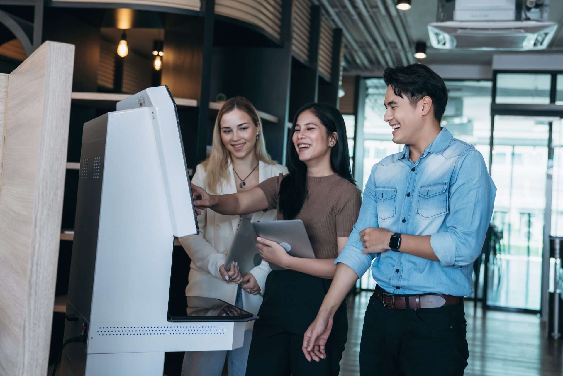 Three smiling people interacting with a modern white self-service kiosk, one pointing at the screen.